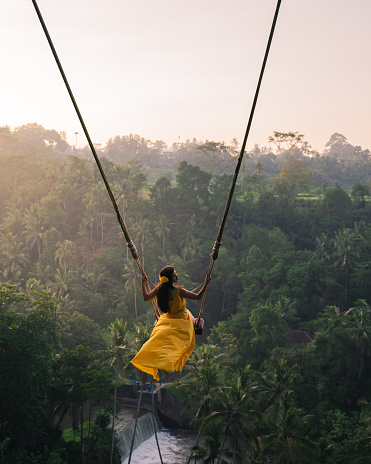 Rear view of a Women sitting on swings at a height of more than twenty meters. In Indonesia Bali Province. Photo of Young tourist woman swinging on the cliff in the jungle rainforest of a tropical Bali island. Young woman swinging in the jungle rainforest of Bali island, Indonesia. Swing in the tropics.