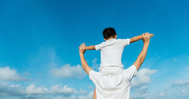 istock Son sitting on father's shoulders against blue sky 1156916822