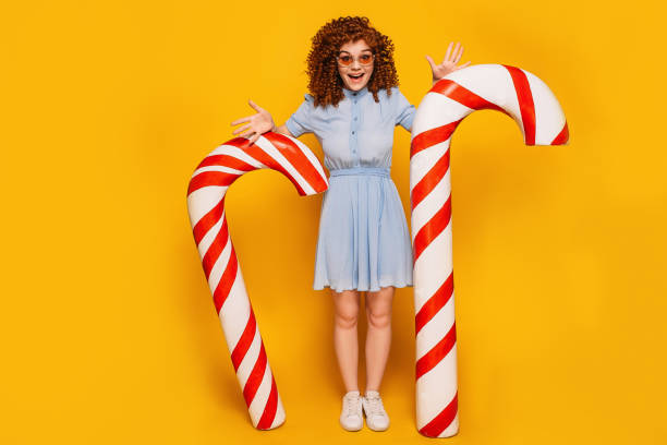 curly red-haired positive woman standing with sweet big candy cane on yellow background curly red-haired positive woman standing with sweet big candy cane on yellow background Candy Cane CANDY stock pictures, royalty-free photos & images