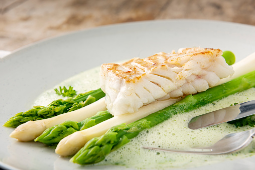 A dish of green and white asparagus topped with a piece of pan fried cod, surrounded by a parsley sabayonne foam. Colour, horizontal with some copy space.