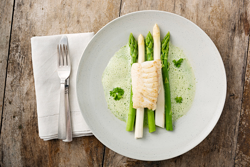 Overhead shot of a dish of green and white asparagus topped with a piece of pan fried cod, surrounded by a parsley sabayonne foam. Photographed against a rustic wooden background, colour, horizontal format with some copy space.