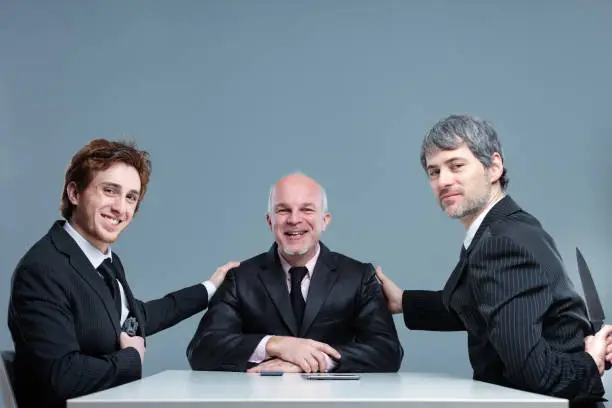 Smiling successful business team posing together with their leader the two young men touching his shoulder and supportive but one holding a knife behind his back conceptual of ambition and betrayal