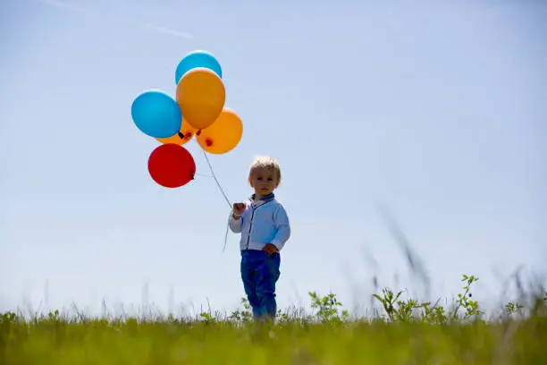 Photo of Little boy, toddler, child playing with colorful balloons in the park on kids day, sunny summer afternoon