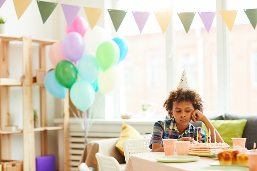 Portrait of sad African-American boy sitting alone at Birthday party in decorated room, copy space