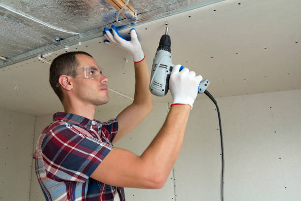 istock Young man in goggles fixing drywall suspended ceiling to metal frame using electrical screwdriver on ceiling insulated with shiny aluminum foil. Renovation, construction, do it yourself concept. 1156942487