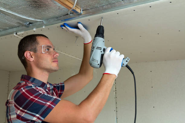 istock Young man in goggles fixing drywall suspended ceiling to metal frame using electrical screwdriver on ceiling insulated with shiny aluminum foil. Renovation, construction, do it yourself concept. 1156942490