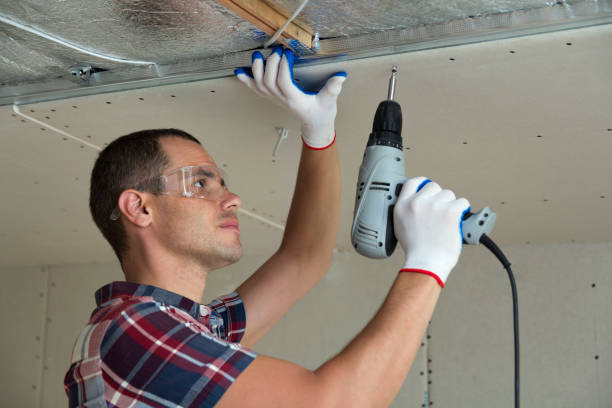 istock Young man in goggles fixing drywall suspended ceiling to metal frame using electrical screwdriver on ceiling insulated with shiny aluminum foil. Renovation, construction, do it yourself concept. 1156942503