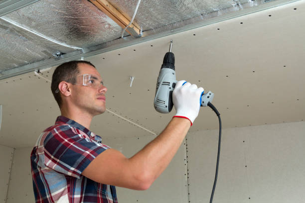 istock Young man in goggles fixing drywall suspended ceiling to metal frame using electrical screwdriver on ceiling insulated with shiny aluminum foil. Renovation, construction, do it yourself concept. 1156942518