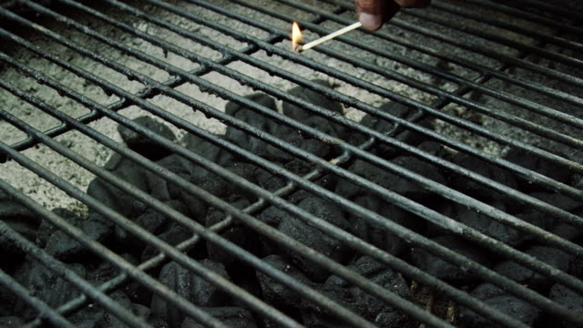 A Caucasian Man's Hand Drops a Lit Wooden Match into a an Outdoor Barbecue Grill, Lighting the Charcoal Briquettes on Fire in Preparation for Grilling