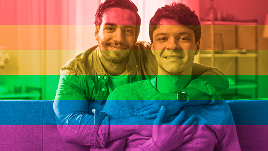 Portrait of a Gentle Male Gay Couple at Home with Rainbow Flag Filter on it. Young Man Sits on a Couch, His Partner Embraces him from Behind. They are Happy and Smiling. Room Has Modern Interior.