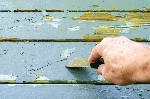 Man's hand shown scraping old gray paint from wood porch boards