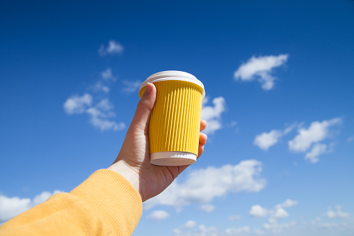 Close-up of female hand holding hot yellow cup of coffee at early morning against the blue sky. Enjoy, lifestyle, take away breakfast concept.
