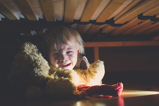 Little child, hiding under the bed, hugging teddy bear and holding flashlight, scared and sad