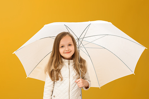 Portrait of a cheerful child in a beige jacket on a yellow background. Little girl blonde stands under the umbrella. Autumn concept.