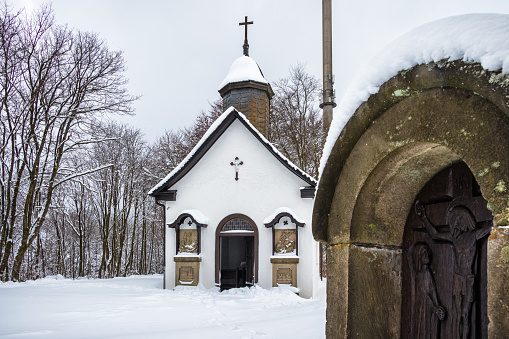Small, snow-covered chapel with prayer stone in the foreground in a winter landscape in Winterberg, Sauerland, Hochsauerland, Germany.
