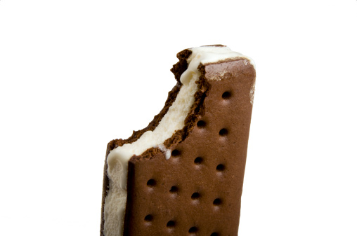 A temptingly delicious ice cream sandwich with a bite already taken and some slight melting. It just makes your crave one.