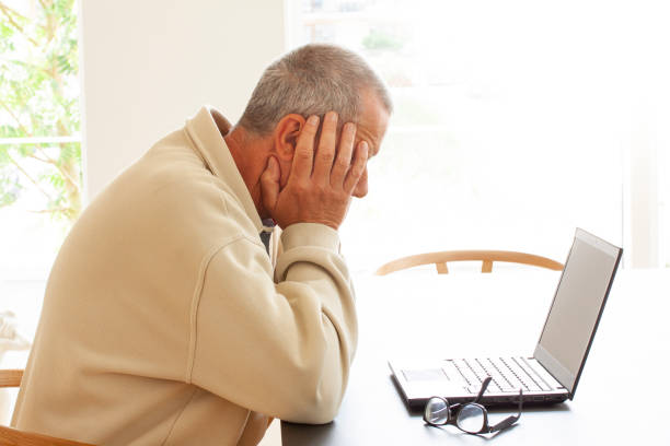 istock Casual dressed man sitting by a laptop computer is hiding his head in despair. A pair of glasses lying on a pile of papers next to the laptop. - Image 1156964511