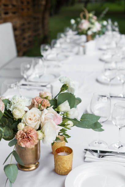 istock Wedding table setting decorated with fresh flowers in a brass vase. Wedding floristry. Banquet table for guests outdoors with a view of green nature. Bouquet with roses, eustoma and eucalyptus leaves. 1156986438
