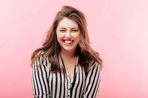 Lovely overweight lady in stylish striped blouse squinting and laughing while standing on pink background