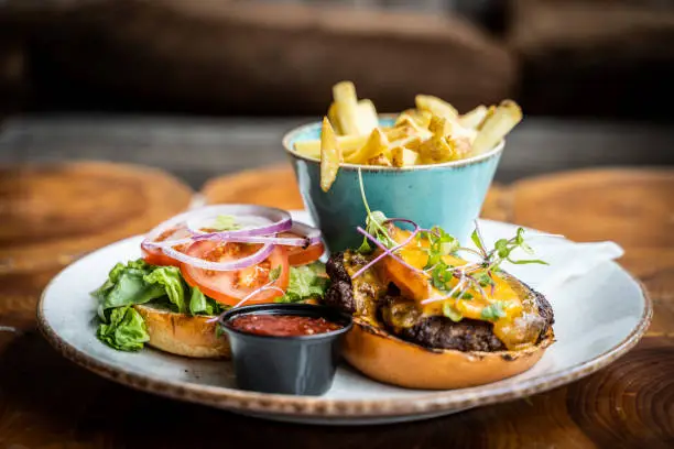 Photo of Cheese burger with bun, fries and sauce on circular plate on a wooden table surface, served in a pub or restaurant with shallow depth of field