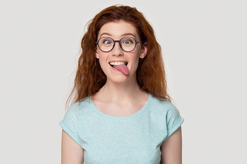 Funny red-haired young girl in round glasses and casual clothes play childish with foolish faces, happy smiling redhead female geek in spectacles isolated on grey background have fun showing tongue