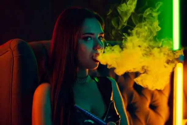 Photo of Beautiful brunette model is smoking a hookah exhaling a smoke at a luxury night club