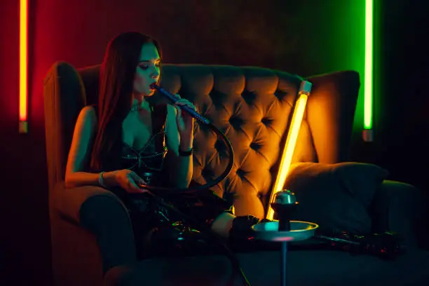 Photo of Beautiful brunette model is smoking a hookah exhaling a smoke at a luxury night club