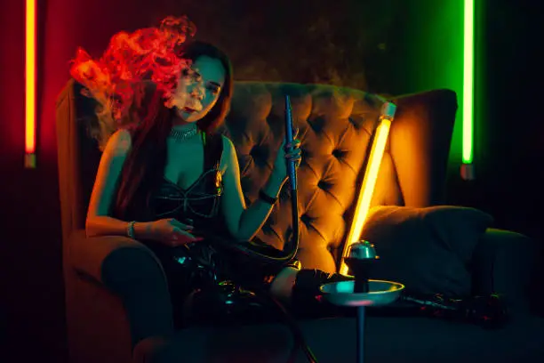 Photo of Beautiful brunette model is smoking a hookah exhaling a smoke at a luxury night club