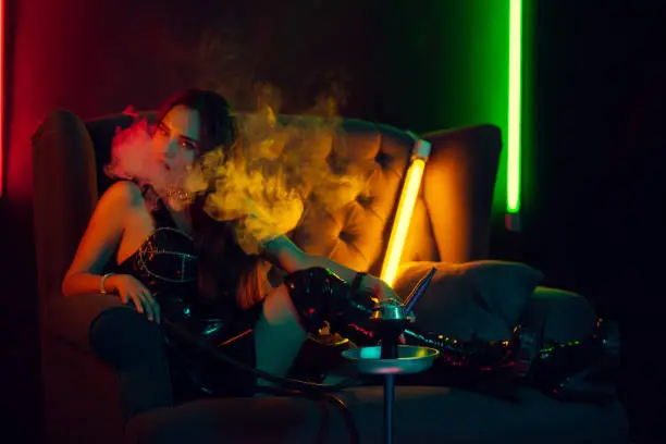 Photo of Beautiful brunette model is smoking a hookah exhaling a smoke at a luxury night club