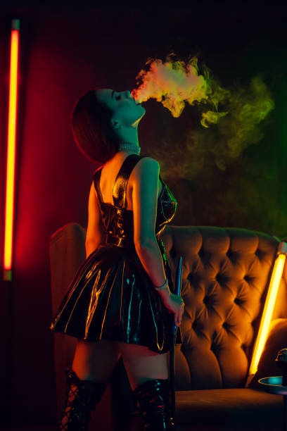 Beautiful brunette model is smoking a hookah exhaling a smoke at a luxury night club Attractive brunette lady with a perfect make-up, in a sparkling necklace, leather dress and patent boots is smoking a hookah exhaling a smoke standing back at a luxury night club. Concept of pleasant spending time. Dark background whith colorful neon backlights. seductive women stock pictures, royalty-free photos & images