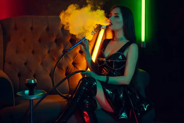 Photo of Beautiful brunette model is smoking a hookah exhaling a smoke at a luxury night club