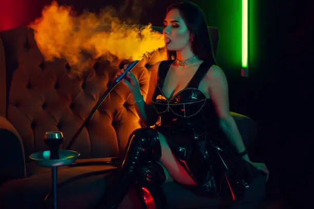 Photo of Beautiful brunette model is smoking a hookah exhaling a smoke at a luxury night club