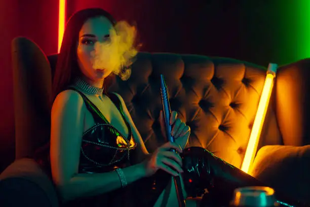 Photo of Beautiful brunette model is smoking a hookah exhaling a smoke at a luxury night club