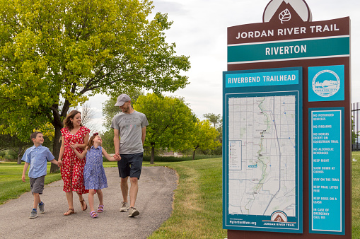 This family is enjoying their time together near the Jordan River Trail in northern Utah. The family consists of mother, father, son and daughter.  Visible in the photo is a sign showing directions for the trail in a public park.  This shot was taken  in Riverton Utah near the entrance to the trail in June of 2019.