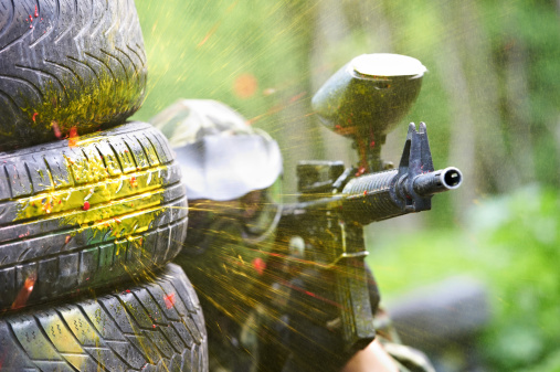 paintball sport player wearing protective mask aiming gun from shelter under gunfire attack with paint splash