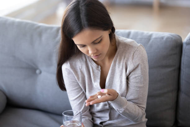 Pensive girl sit on couch doubt to take pills Pensive girl sit on couch holding medicines and glass of water thinking of taking medications, depressed young woman on sofa pondering doubt whether to take pills antidepressant or contraceptives headache menstruation pain cramp stock pictures, royalty-free photos & images