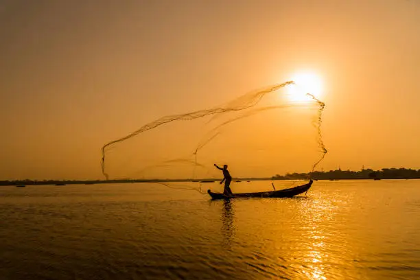 Silhouette Asian fisherman on wooden boat casting a net for freshwater fish at U Bein Bridge,Mandalay, Myanmar (Burma) in the early morning before sunrise