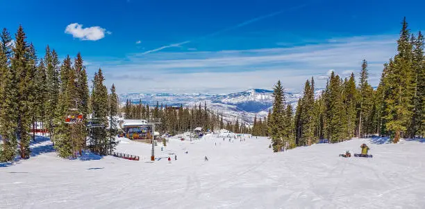 View of chairlift and ski slope in Colorado ski resort with Colorado spruces on both sides and mountains in background; skiers and snowboarders on slope