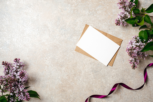 Flat lay of kraft envelope and blank paper card with copy space mock up. Top view home office desk workspace decorated with lilac flowers and purple ribbon. Beauty blogger, minimal women desk concept.