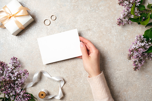 Hand of woman holding blank paper card over feminine desk with wedding accessories and spring lilac flowers. Wedding concept, invitation card mock up, flat lay, top view.