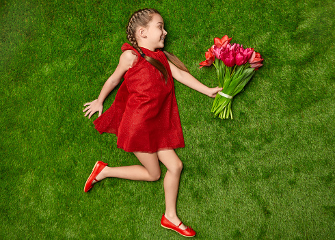 Side view of cute little girl in red dress smiling and holding bunch of beautiful flowers while lying on green grass