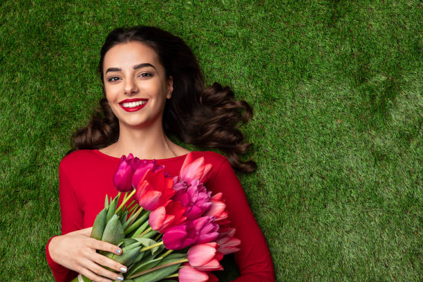 Overhead view of charming brunette in red dress and with red lips lying on green grass with bright tulips. Women day concept with copy space