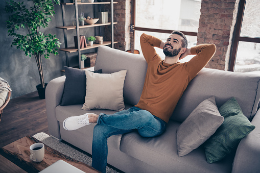 Portrait of his he nice attractive bearded cheerful cheery content guy sitting on divan having rest at industrial loft interior wooden brick style living-room, indoors