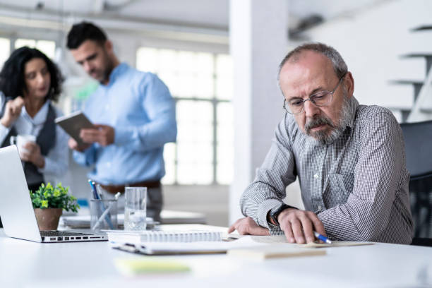 istock Senior businessman using laptop, behind coworkers talking 1157094407