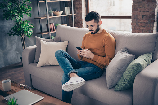 Portrait of his he nice attractive focused concentrated bearded guy sitting on divan surfing, web addict at industrial loft brick interior style living-room