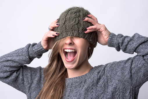 Portrait of screaming young woman with knitted cap covering her eyes on light gray background.