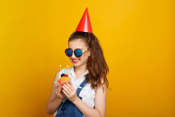 Portrait of a happy young girl in sunglasses with bright makeup over yellow background, holding in hands a cupcake with candles, dressed in summer clothes, on head have a red cap. stock photo