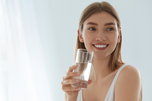 Woman with glass of fresh water in white portrait. Beautiful smiling girl with healthy skin and beauty face holding pure mineral water in glass indoors