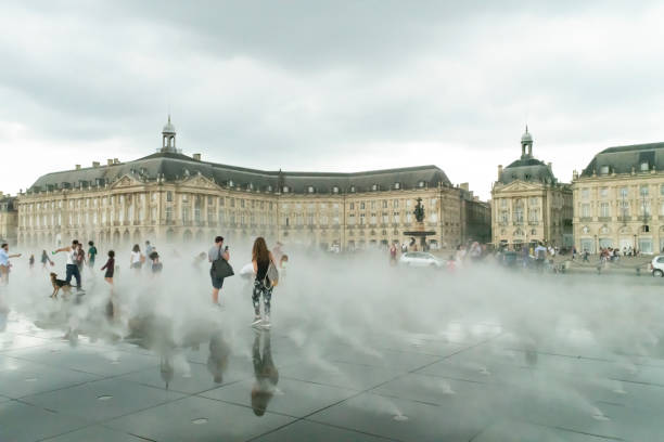 Place de la Bourse at Bordeaux, France this is a Unesco World Heritage site dating from the 18th century. There are many tourists walking in the foreground across a pavement made up of water jets which spray alternatively water and mist to create a fog like appearance.  In the background is the Bourse or stock market which is the main element of the UNESCO site. It was built from 1730 to 1775 by architect Ange-Jacques Gabriel, it is neither a castle or palace.