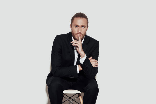 istock Keep my secret! Handsome young man in full suit looking at camera and keeping finger on lips while sitting against grey background 1157118922